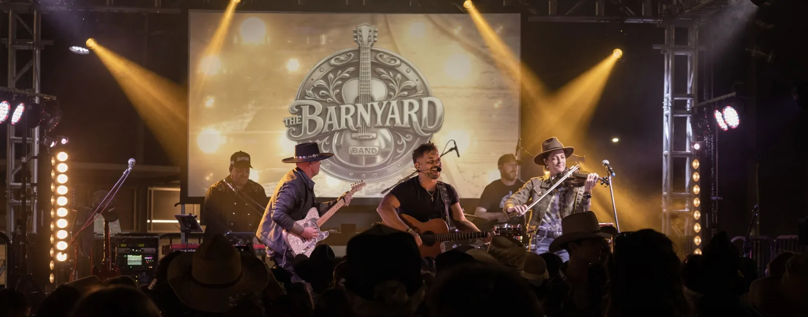 The Barnyard Band live on stage under gold spotlights, with the Barnyard logo on the big screen and the crowd's hands in the air